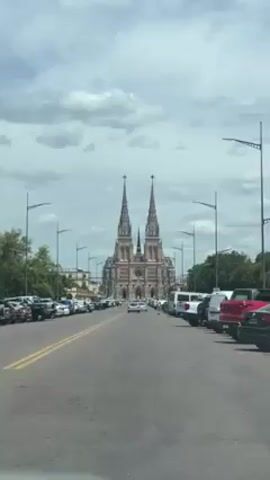 The camera, from inside a moving vehicle, captures a view down a street leading to a large cathedral with two prominent spires. The street is lined with parked cars on both sides, and there are a few vehicles moving in the same direction as the camera. The sky is overcast with a mix of clouds. There are streetlights lining the road on both sides. The cathedral is the focal point, its architecture detailed despite the distance. In the audio, there is music playing with a strong rhythm.