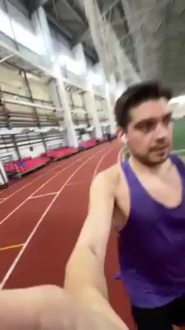 A young man jogs on a red indoor running track while filming himself with a handheld device. He wears a purple tank top and black shorts, with earbuds in his ears and an orange band on his wrist. The camera angle is low and points slightly upward. The indoor track has white lines and is bordered by green turf. Some spectators are visible in the background. He jogs past starting blocks as the video ends.