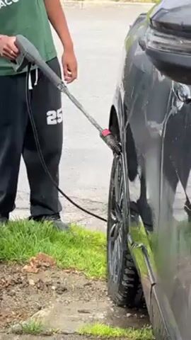 A young man is shown cleaning a dark-colored car with a pressure washer. He is wearing a green Lacoste t-shirt, black sweatpants with "25" on the leg, and black shoes. He is standing on a patch of grass next to a paved road. The car is parked alongside a metal gate. The background shows a residential area with small houses and a distant mountainous landscape. The man starts by spraying the car's wheel well and then moves to clean the car body, standing behind the gate. The shot slightly zooms in on the man's face as he cleans the car.