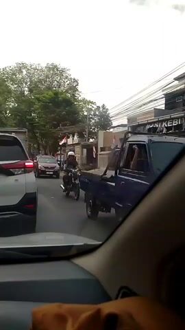 A person on a motorcycle wearing a red helmet and a light-colored shirt rides down a paved road. They are passing a maroon car in the lane to their left. The motorcycle is dark-colored with some chrome accents. In the background, there are buildings on the right side of the road, one of which has signage that appears to say "NASI KEBULI." There are also trees and utility poles visible. A white car is in the left lane, partially visible in the foreground, and a person is visible in the driver's seat. The sky is overcast.