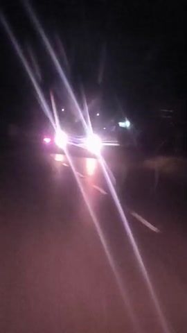 The video captures a dark, nighttime road scene, viewed from a moving vehicle. Bright white headlights of oncoming vehicles create prominent streaks of light across the frame, indicating motion. Red taillights of other vehicles are also visible further down the road, appearing as blurred points of light. The road surface is visible in the foreground. The overall setting suggests driving at night on a road, possibly in a rural or semi-urban area due to the lack of extensive artificial lighting in the background. No people or visible text are discernible in the footage. The provided location context is Gampongrayadagang, Indonesia.