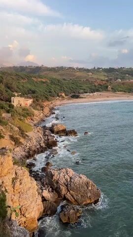 The video opens with a shot of a rocky coastline with turquoise water and waves crashing against the rocks. In the distance, there is a beach with some buildings and vegetation. The camera then pans to the right, showing more of the coastline and the ocean. The sky is cloudy with hints of pink and orange, suggesting either sunrise or sunset. The video continues to pan across the ocean, revealing a vast expanse of water under a dramatic sky. The sun is visible on the horizon, casting a warm glow over the scene. The video ends with a shot of the sun setting over the ocean, with clouds reflecting the golden light.