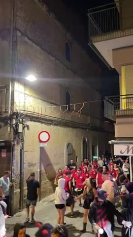 A group of people are gathered in a narrow street at night. Many are wearing red shirts and some are wearing black and red costumes. Some individuals are holding objects that appear to be drums or sticks. There is a significant amount of activity and movement within the crowd. The street is lined with buildings, one of which has a large, textured wall. There is a street light illuminating part of the scene. A street sign with a red circle and a white bar is visible on a pole. Decorations, possibly strings of objects, are hung across the street from the buildings. The atmosphere appears to be festive, with people dancing and interacting. The ground is paved, and the overall lighting suggests it is nighttime.