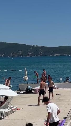 A group of boys are playing on a sunny day at the beach. The boys are wearing swim trunks and are barefoot. Some of them are jumping into the water from a concrete ledge. Others are standing on the ledge or on the beach, watching or playing with a soccer ball. A few beach umbrellas and lounge chairs are visible in the foreground. The water is a clear turquoise color, and a line of buoys separates the swimming area from the open sea. In the background, a green, tree-covered hill slopes down to the water. The sky is a clear blue.