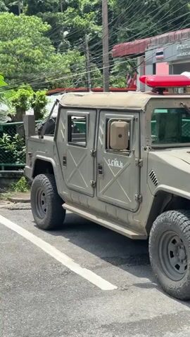 The video shows a close-up, eye-level side view of a light olive-drab military-style vehicle parked on an asphalt road. The vehicle appears to be a Humvee or similar high-mobility multi-purpose wheeled vehicle. It has a canvas top and four doors, with the front passenger door and rear passenger door visible. There is writing in an unknown script on the front passenger door. The vehicle is equipped with a light bar on the roof, featuring red and blue lights. Large, rugged tires are visible on both the front and rear axles. The front of the vehicle, including the grille and headlights, is partially visible on the right side of the frame. Behind the vehicle, a building with a red awning and a sign with Thai writing is visible, along with trees and power lines. The overall impression is that the vehicle may be a police or emergency response vehicle in a tropical or sub-tropical location. The lighting suggests it is daytime.