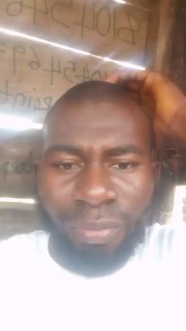 A close-up shot shows a man with dark skin, a beard, and a shaved head wearing a white shirt. He appears to be speaking to the camera, and his facial expressions change. His right hand is visible on the top of his head, as if he is pushing his head down. He is indoors with weathered wooden walls behind him. Reversed writing is also visible on the wall. The light is bright, but the image is slightly blurry.
