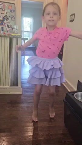 A young girl with long brown hair, tied back with a pink ribbon, is dancing in a room with wooden floors. She is wearing a pink t-shirt with small black hearts and a light purple, tiered, ruffled skirt. She has bare feet. The background shows a kitchen area with cabinets and a refrigerator adorned with magnets and photos. A doorway leads to another room, where a window is visible. To the right, a dark object with a silver bowl on top is present. The lighting suggests it is daytime. The girl appears energetic and joyful as she moves.