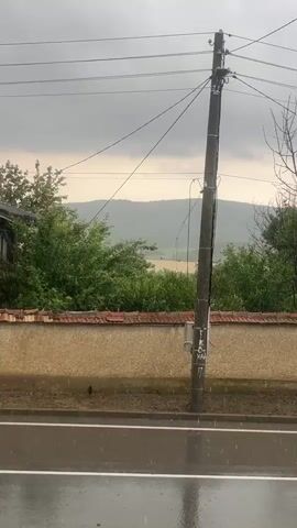 A fixed outdoor shot shows a rainy, overcast day. Raindrops fall on a wet asphalt road in the foreground. To the right of the road is a dirt strip with small plants, and then a low tan wall topped with terracotta tiles. Behind the wall, green trees and bushes are visible. A tall, dark utility pole stands at the center-right of the frame, with multiple black wires extending horizontally across the upper portion of the frame. In the background, rolling green hills are visible under a grey, cloudy sky. No people or animals are present.