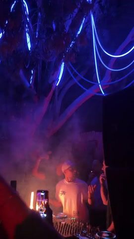 The video shows a DJ performing at a club. The DJ, wearing a white t-shirt and a cap, is standing behind a DJ console. Several people are in front of the DJ, dancing and holding up their phones to record the performance. The club is dark, with blue and red lighting, and smoke fills the air.  Some people are wearing black clothing. The DJ has headphones around their neck and they are touching the controls.