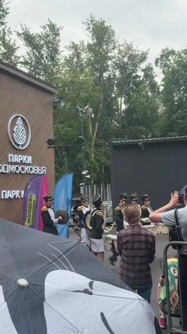 This is a vertical, eye-level video capturing an outdoor event. The primary subjects are a marching band of young people dressed in military-style green and gold uniforms with black hats. They are playing drums and cymbals, suggesting a performance or parade.

The setting appears to be a park, with a building featuring a logo and text visible on the left. Trees and foliage form a backdrop, indicating a natural or semi-natural environment. The sky is overcast, and raindrops are visible, along with a black umbrella with a white cat design in the foreground, suggesting it is raining or has recently rained.

The text on the building reads "ПАРКИ ПОДМОСКОВЬЯ" (Parks of the Moscow Region) and "ПАРК М" (Park M). Two flags, one purple and one blue, are also visible, with text on them that is partially obscured but appears to include "МОЛОДЕЖЬ" (Youth) on the purple flag.

There are several spectators visible, some holding umbrellas, and one person in the foreground is recording the event with a phone. The overall mood seems festive and engaged, despite the weather. The time of day is indeterminate due to the overcast conditions, but it appears to be daytime. The location context is Kolomna, Russia.
