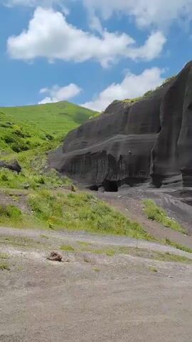 A silver SUV is parked in a gravelly area at the base of a large, dark, rocky cliff face. The cliff has distinct horizontal layers and vertical striations carved into it, possibly by wind or water erosion. A cave-like opening is visible in the cliff face. Green grass and small yellow flowers grow on a sloped area to the left of the cliff. The sky above is blue with scattered white clouds. The camera angle is slightly low, looking up at the cliff and the vehicle.