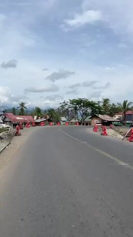The video shows a paved road with yellow lane markings. The road curves to the right, and on either side are several red and white traffic barriers. Beyond the barriers, there are buildings, some with corrugated metal roofs, and trees, including palm trees. The sky is cloudy. The video appears to be taken from a moving vehicle. There are no people visible in the video.