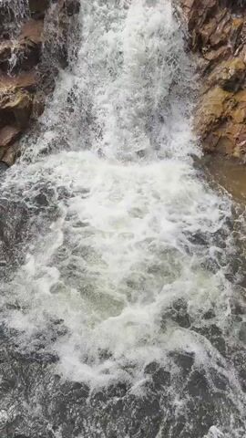 THE VIDEO SHOWS A CLOSE-UP OF A WATERFALL DESCENDING OVER RUGGED ROCKS. THE WATER IS WHITE AND FOAMY, CREATING A TURBULENT EFFECT AT THE BASE. THE ROCKS ON EITHER SIDE OF THE WATERFALL ARE BROWN AND CRAGGED, WITH SOME GREEN VEGETATION VISIBLE AT THE TOP OF THE FRAME. THE CAMERA APPEARS TO BE POSITIONED DIRECTLY IN FRONT OF THE FALLING WATER, OFFERING A VERTICAL PERSPECTIVE. THE FOOTAGE IS SHAKY AT TIMES, SUGGESTING IT WAS FILMED HANDHELD.