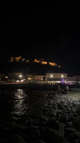 The video shows a nighttime scene on a rocky beach. In the foreground, dark rocks are piled up. The water of a body of water, likely the sea, is visible on the left, reflecting the lights from the shore. Along the shore, there are several rows of beach umbrellas, most of which appear to be closed or folded. Behind the umbrellas, there are buildings, illuminated by yellow and white lights. Further in the background, a large, ancient-looking castle or fortress is situated on a hill, also illuminated by lights, casting a warm glow against the dark night sky. A few people are visible on the beach near the rocks in the right foreground, and some more are seated at tables under a canopy-like structure behind the umbrellas, suggesting a restaurant or bar. The overall atmosphere is peaceful and inviting.