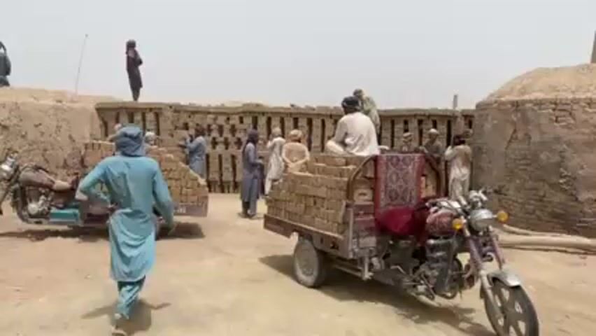 The video depicts a group of people in a dusty, seemingly rural area.  They appear to be working at a brick kiln or brick-making facility.  Several men are loading or have loaded bricks onto small three-wheeled vehicles attached to motorcycles.  One man in a white outfit is sitting atop a load of bricks on one of the motorcycle-trikes.  The environment includes brick structures and a relatively flat, open space.  The sky appears overcast. The people are dressed in various traditional outfits.