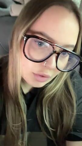 A young woman with long, multi-toned (blonde and brown) hair is shown from a close-up, slightly low angle. She wears large, dark-framed glasses with a prominent top bar and has a small hoop piercing in her left nostril. She is wearing a black shirt. Throughout the video, she moves her head slightly, looking forward and occasionally to her left, and her mouth is open and moving as if she is speaking or singing. The background appears to be the interior of a vehicle, possibly a car, with a headrest visible behind her. Ambient music is audible.