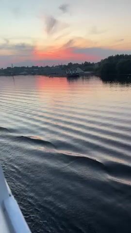 A video captures the surface of dark, rippling water, likely a river or lake, with waves extending towards the viewer. In the distance, a hazy shoreline is visible, featuring a few structures and trees. A small boat with a white cabin and red roof is anchored near the shore. The sky above is painted with the soft colors of a sunset, with hues of pink, orange, and yellow blending with light gray clouds. The reflection of the sunset is visible on the water's surface. The perspective suggests the video is taken from a moving boat.