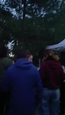 A group of about 20 people are standing in an outdoor setting. Some are interacting with each other, hugging, talking, and high-fiving. It appears to be dusk, with trees visible in the background. There's music playing, and the atmosphere seems festive. Individuals are dressed in casual clothing like jackets, jeans, and sweaters.