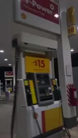 The video shows a deserted Shell gas station at night. The station is brightly lit by overhead lights and features a red and yellow trim along the roof of the canopy. There are multiple gas pumps, with their price displays visible. No people or cars are visible, creating an atmosphere of solitude.