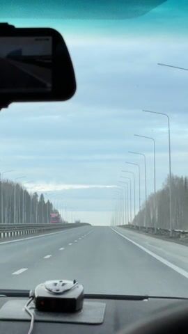 The video shows a view from inside a car driving on a highway. The sky is overcast, with tall streetlights lining the side of the road. A red truck is visible in the distance on the left side of the road. The road is multi-lane and appears to be in good condition. A radar detector is visible on the dashboard.
