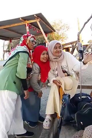 This is a video of three women posing for a selfie at what appears to be an outdoor market or gathering. The woman on the left is wearing a green hijab, sunglasses, and striped pants. The woman in the middle is wearing a red hijab and a gray jacket. The woman on the right is wearing a light pink hijab and a white t-shirt. She is holding a selfie stick with a phone attached. The women are smiling and laughing, and the woman in the middle is pointing towards the camera. There are bags and other items on the ground around them. The background shows a wooden structure with a canopy and some trees.