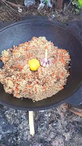 A top-down shot shows a large, black, cast-iron cooking pot on a makeshift grill over a dying fire.  The pot contains a mound of rice, cubed carrots, and what appear to be pieces of meat, all mixed together.  Nestled on top of the rice mound are a whole yellow lemon and a full head of garlic. Some steam is rising from the rice. The surrounding area appears to be a rustic, outdoor setting, possibly in a rural or natural environment, with scattered vegetation and debris. The camera position is steady.