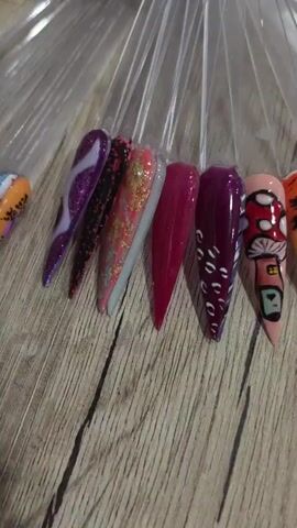 The video displays a collection of artificial nails, each attached to a plastic tip and fanned out on a wooden surface. The nails exhibit a variety of designs and colors, including glitter, abstract patterns, and painted images such as mushrooms, palm trees, and a flamingo. The lighting suggests an indoor setting. The camera pans across the nails, offering a close-up view of each design.