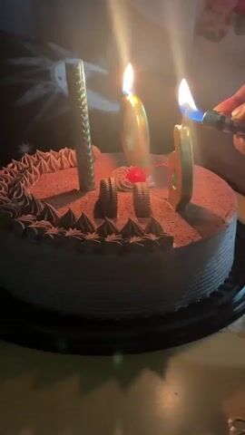 A close-up shot shows a chocolate cake adorned with chocolate frosting, chocolate shavings, two Oreo cookies, a red cherry, a silver sparkler-like candle, and two golden numeric candles shaped as "6" and "0". A hand from the right uses a blue-flamed lighter to ignite the "0" candle, then moves to light the "6" candle. Both candles are already lit at the start of the clip. The cake rests on a black cake board against a dark background with faint, illuminated patterns.