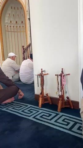 The video shows a group of men sitting on a blue carpet in a mosque. The men are facing a wall, with some in prayer positions and others seated. The mosque has ornate architectural features, including arched doorways and intricate wall decorations. Two wooden stands with prayer beads are visible in the foreground. The lighting suggests it is daytime.