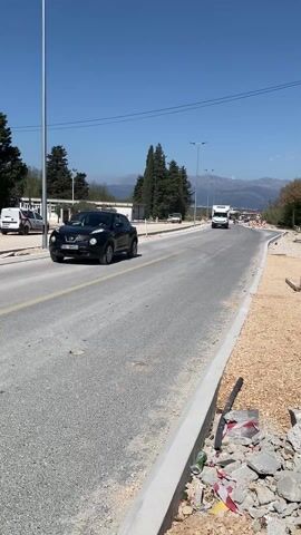 The video shows a road construction site. A black car is parked on the left side of the road. A white truck drives on the road followed by a dump truck. Debris is on the road’s shoulder on the right side. The sky is clear and blue. In the background, there are trees and snow-capped mountains.