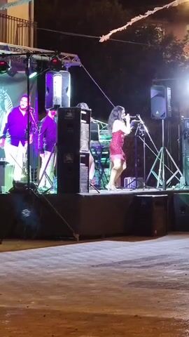 The video shows a live musical performance outdoors at night. A band is on a stage equipped with lighting and speakers. On the left side of the stage, two men are visible; one appears to be playing an instrument, and the other is standing. In the center of the stage, a woman in a red sequined dress is singing into a microphone and dancing. Another woman in a similar red dress is standing behind her, also holding a microphone. The stage is set up on a paved area, possibly a street or plaza, with some buildings and trees in the background, illuminated by stage lights and some overhead string lights. The overall atmosphere suggests a festive outdoor event.