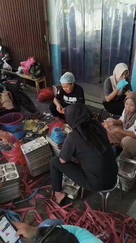 The video shows a group of four women preparing food outdoors. They are seated on the ground amidst piles of aluminum food containers and pink plastic strapping. The women are wearing head coverings, with two wearing hairnets and two wearing hijabs. One woman in the background is wearing a black t-shirt with white writing on it. They are all engaged in handling food items and the containers. The environment appears to be a covered outdoor area, possibly a loading dock or a preparation space adjacent to a building. Motorcycles are parked behind the women. One person in the foreground is holding a smartphone, looking at the screen. The overall scene suggests a food preparation or packaging operation.