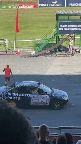 The video shows a car stunt performance at an outdoor venue, likely a racecourse, given the grassy field and grandstands visible in the background. In the foreground, spectators are seated in a covered grandstand, some holding up phones to record. Two cars are on a paved area. One is a dark-colored car, possibly made to resemble a police car with "POLICE" scrawled on its side in red paint and yellow tape along the top. It has a blue flashing light on its roof. The other car is red and has "IRISH AUTO PARTS" written on its side with a circular logo. Both cars appear to be participating in a stunt, as evidenced by tire marks on the pavement and the way the cars are positioned. In the background, there's a ramp structure leading up to a platform with a sign that reads "THE TEAM AT PUNCHESTOWN WISH YOU AN ENJOYABLE DAY." There are also several advertising banners visible, including "LADBROKES" and "PUNCHESTOWN." The weather appears to be sunny with clear skies.