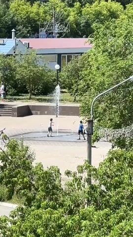 In a sun-drenched plaza, three young boys play in a fountain. One boy in a light-colored shirt and dark shorts runs towards the fountain. Another boy, wearing a dark shirt and shorts, stands in the fountain. A third boy, in a blue shirt and dark shorts, also stands in the fountain, facing the other two. The fountain sprays water upward, and the boys appear to be interacting with the water. The plaza is paved with light-colored tiles, and a few dark square outlines are visible in the fountain area. Trees with lush green foliage surround the plaza. In the background, a building with a blue roof and a red roof is visible, along with more trees. A lamppost stands to the right of the frame.