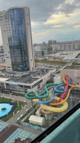 This media file provides an aerial view of an outdoor water park complex situated in Kzyl-Bairak, Russia. The scene captures a large, modern water park with multiple attractions, including vibrant, colorful water slides in yellow, blue, green, and red, spiraling into a pool area. Adjacent to the slides are several irregularly shaped swimming pools, some with lounge chairs and umbrellas set up around them, suggesting a recreational setting.

A prominent feature of the landscape is a tall, modern building with a glass facade, likely a hotel or a commercial establishment, towering over the water park. Beyond the immediate water park amenities, the view extends to a construction site with bare earth and scaffolding, indicating ongoing development in the area. Further in the background, a cityscape unfolds with numerous residential buildings and urban infrastructure. The edge of a body of water, possibly a river or lake, is visible on the left side of the frame, with a long, rectangular swimming pool labeled "РИВЬЕРА" (Riviera) alongside it.

The time of day appears to be either sunrise or sunset, indicated by the soft, diffused light and the colorful hues in the sky, with clouds adding texture to the atmosphere. The overall mood conveyed is one of leisure and a bustling, growing urban environment. There are no people explicitly visible in the frame, but the presence of lounge chairs and the design of the water park strongly suggest that this is a place for public enjoyment and activity. The combination of modern architecture, recreational facilities, and ongoing urban development paints a picture of a dynamic and developing location.