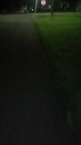 A first-person perspective video begins with a nighttime view of a paved path or road with grass on the right. Light streaks are visible in the distance. The view then transitions to darkness. Suddenly, a person's face and shoulder wearing a plaid shirt come into view, their face partially obscured and out of focus. The scene shifts again to a dark environment with a distant light source that appears to be a streetlamp. The view then moves to a close-up of a person wearing a plaid shirt and a gray hooded sweatshirt or blanket.
