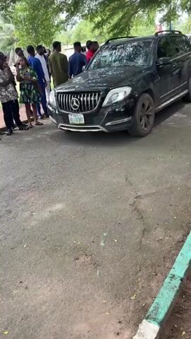 The video shows a black Mercedes-Benz SUV parked on an asphalt road next to a curb painted with alternating green and white sections. A group of people are standing in the background, some holding phones. The SUV has a license plate visible. The road is littered with small debris. The camera moves from the SUV towards the curb.