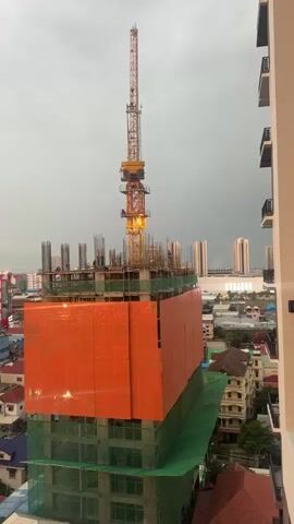 The video shows a construction site in a city under a cloudy sky. A tall crane extends from the top of an unfinished building. The building is partially covered in orange netting and green scaffolding. Workers can be seen on the upper levels of the construction site. In the background, other buildings and a hazy horizon are visible.
