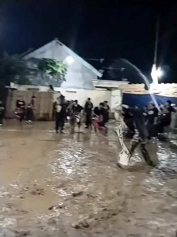 The video captures a nighttime scene in Jenggrik, Indonesia, where a group of people are participating in a cultural or traditional event on an extremely muddy ground. Many participants are dressed in dark clothing, some with traditional elements like patterned fabric around their waists or legs, while others wear casual attire such as t-shirts, shorts, and caps.

In the foreground, a person covered in mud is seen holding a white cloth or flag. Another individual, also covered in mud, is on all fours wearing a white headpiece or mask, moving in a manner suggestive of an animal or a trance state. Two muddy horses are partially visible on the right side of the frame.

The setting includes buildings in the background, a street light illuminating parts of the scene, and a red and white flag (likely Indonesian) flying on a pole. A blue tarp and wooden structures are also visible in the background. The event appears energetic, with people moving and splashing in the mud. The combination of traditional dress, trance-like performance, mud, and horses strongly suggests a "Jaranan" or "Kuda Lumping" performance, a traditional Javanese dance. No visible text is clearly legible in the video.