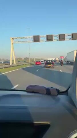 The video shows a view from the inside of a car driving on a highway. There are multiple lanes of traffic with various vehicles including cars, a motorcycle, and a semi-truck. Overhead, there is a road sign structure with several blank rectangular signs. The weather is sunny with a clear blue sky. The car's dashboard is visible, with a gray and black cloth on top.