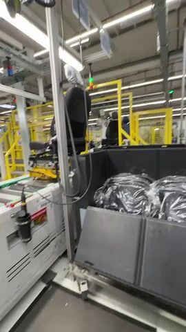 The video shows a factory assembly line with car seats being manufactured. In the foreground, a car seat is mounted on a movable frame. To the right, a container holds several car seats wrapped in plastic. The assembly line features a conveyor belt system, robotic arms, and control panels. Yellow safety barriers are visible throughout the factory. The environment appears to be a well-lit industrial setting with overhead lighting and exposed electrical wiring. The overall scene depicts the automated production of automotive seating.
