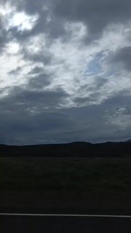 The video shows a view from a moving vehicle, likely a car, looking out a window. The scene is dominated by a dramatic sky filled with dark, heavy clouds, with patches of lighter sky visible. The horizon line shows a landscape of rolling hills or mountains, which are silhouetted against the sky. In the foreground, a dark road with a white line is visible, indicating the vehicle is traveling on a paved road. A field of green vegetation, possibly grass or low shrubs, stretches across the landscape between the road and the hills. There are no people or distinct objects visible in the frame, and the focus is on the natural environment and the atmospheric conditions. The overall impression is one of a vast, somewhat ominous natural landscape under a cloudy sky.