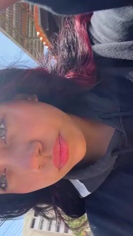 This is a close-up, vertical, low-angle selfie video of a young woman with dark hair, partially dyed with magenta streaks. She is wearing a black hooded sweatshirt. The background appears to be outdoors, with greenery and a glimpse of a building structure suggestive of a balcony or patio, possibly in a tropical setting. The lighting indicates it is daytime, with strong sunlight casting shadows. The woman's face is the primary focus. She is looking directly at the camera with a slight smile and her eyes are open. She has prominent makeup, including bold lipstick in a pink-red shade and eye makeup. There is no visible text in the video. The overall mood is casual and relaxed, possibly captured during an outdoor leisure moment. The setting hints at a warm climate.