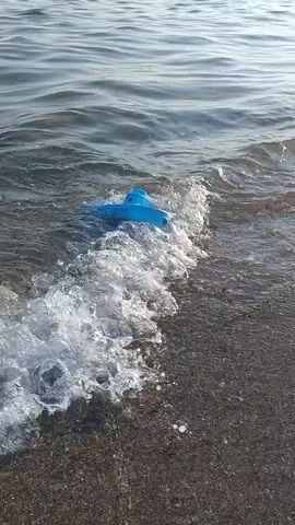 A blue plastic toy boat is being tossed around in the shallow waves at the edge of a beach. The water is somewhat choppy with white foam from the waves. The background shows a body of water stretching to the horizon with a structure visible in the distance. The camera is positioned low, near the water's surface, and is tilted downwards.