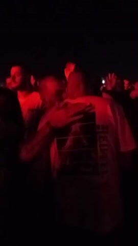 A crowd of people is gathered outdoors at night. Red lighting illuminates the scene, casting a vibrant glow on the attendees. Some individuals are dancing and raising their arms in the air, indicative of a lively atmosphere. Others are conversing and holding drinks, suggesting a social event. The background features tents and structures, possibly indicating an outdoor festival or concert venue. The overall impression is of a vibrant and energetic gathering.