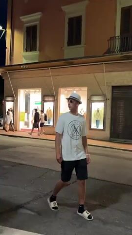 The video shows a group of people standing outside a building at night. The building is light-colored and has windows with closed shutters. Some of the people are standing in a line, while others are standing around them. A few people are wearing black shirts, while one person is wearing a pink shirt and another is wearing a white shirt and a white hat. There is a bush to the right of the group. A store with lighted displays can be seen in the background. A streetlamp illuminates the sidewalk.