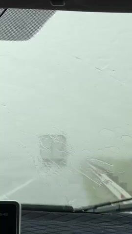 The video shows a view from inside a vehicle looking out through a rain-streaked windshield. The scene outside is very foggy and it's difficult to make out many details. In the distance, a blurry, rectangular shape that might be a building with a window is visible. There's also a suggestion of a red and white striped object on the right side of the frame, possibly a barrier or sign. The overall impression is of a very overcast and wet day, perhaps during a rain shower. The light suggests it is daytime, though the heavy fog makes it difficult to ascertain the exact time. The interior of the car is partially visible at the bottom of the frame, with a device displaying "3:43" which could indicate the time. The texture of the car's dashboard is also visible. The weather is clearly rainy and foggy, obscuring the view of the outside world. The setting appears to be a road or pathway, given the context of being inside a vehicle. The emotional tone conveyed is one of subdued visibility and possibly a sense of being isolated by the weather. There are no identifiable people or animals visible.