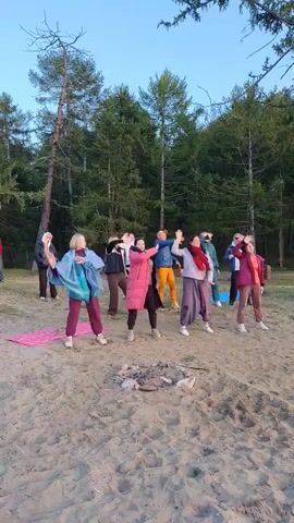 A group of eleven people are gathered on a sandy area, with a forest in the background. The sky is a light blue, suggesting it is either early morning or late evening. The people appear to be engaged in a group activity, possibly dancing or a form of exercise. Some individuals are wearing casual clothing, including jackets and scarves, and the overall atmosphere seems relaxed and informal. One person in the center is playing a guitar, and another person is standing near a microphone stand. In the foreground, there is evidence of a campfire.