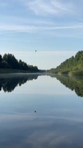 A video begins with a shot of a calm river reflecting the sky and trees on its banks. The camera is positioned low, giving the impression of being on the water. As the video progresses, the view shifts slightly to show the front of a small, weathered boat. A person wearing a bright yellow jacket and dark pants stands in the boat, their back to the camera. They appear to be looking out at the river. The scene is tranquil, with a bright blue sky with scattered white clouds above the tree-lined riverbanks.