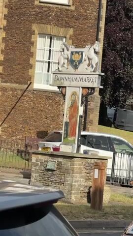 A static medium shot shows a stone-fronted building with a window and a large sign in the foreground. The sign features two horses on top, a crest, and the words "Downham Market." Below, a religious figure is painted on a tall, narrow panel. A white car's roof and spoiler are visible in the bottom left, partially obscuring the bottom of the frame. In the background, a white car is parked behind a metal fence, and a dark van is further back.
